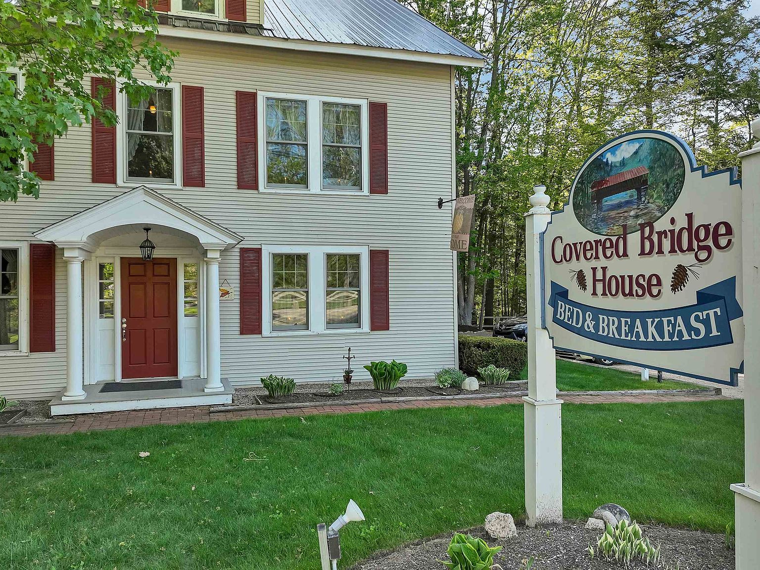 Front facade of the Covered Bridge House Bed & Breakfast, a white clapboard New Hampshire farmhouse with deep red shutters and a hand-painted hanging sign at the lawn
