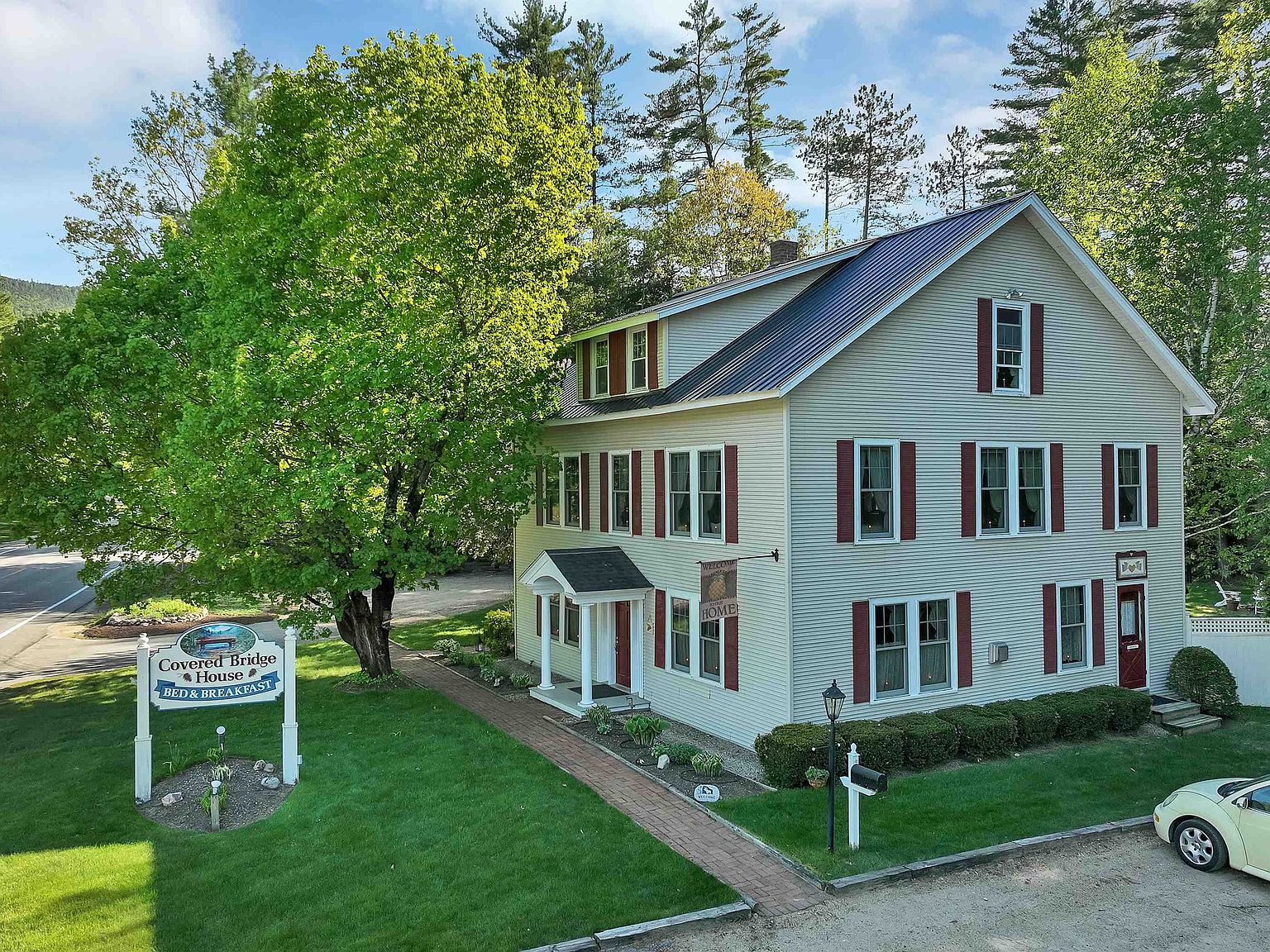 Elevated view of the Covered Bridge House inn from above, showing the standing-seam metal roof, the maple tree in summer leaf at the front lawn, and Route 302 below