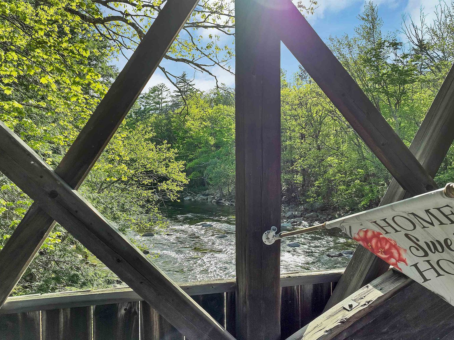 Looking out through the wooden lattice trusses of the covered bridge to the rushing Saco River and sunlit foliage beyond, with a corner of the Home & Bridge Gift Shoppe sign at the right edge