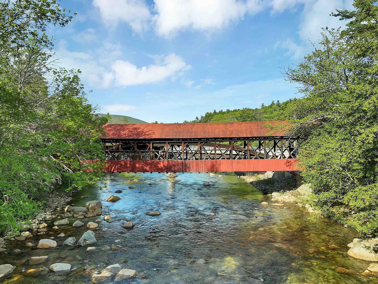 The Covered Bridge House bridge spanning the Saco River, its weathered red Paddleford-truss roof set against summer foliage and the White Mountains beyond
